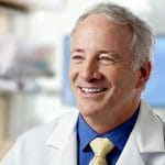 Smiling male doctor in a white coat with a yellow tie and blue shirt, sitting in a bright medical office.