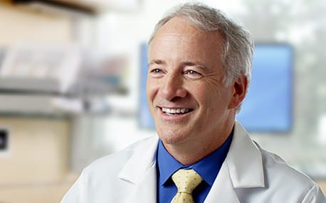 Smiling male doctor in a white coat with a yellow tie and blue shirt, sitting in a bright medical office.