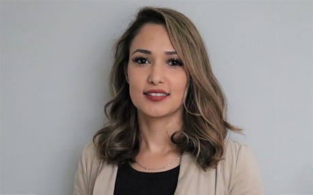 Smiling woman with shoulder-length wavy hair, wearing a light blazer over a black top, posed against a plain gray background