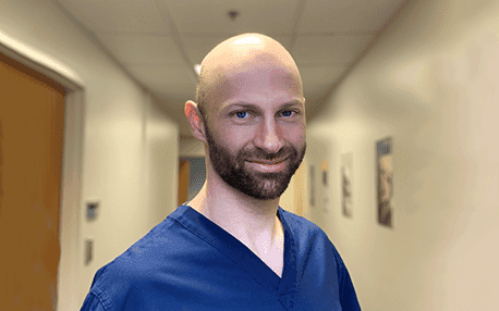Man with a bald head and beard wearing navy blue medical scrubs, standing in a well-lit hallway with framed artwork.