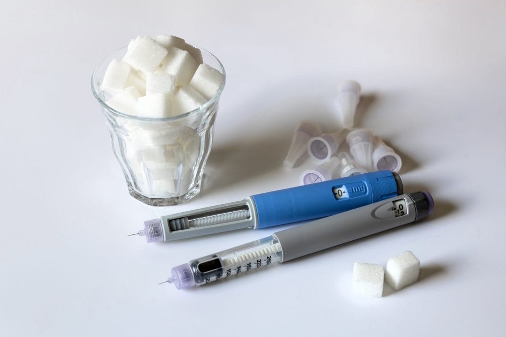 Two semaglutide injection pens lying next to a glass filled with sugar cubes and several needle caps, symbolizing treatment for obesity.