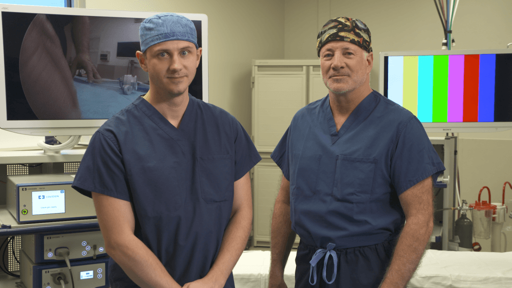 Dr. Vanderwel and Dr. Landerholm in front of an Operating Table wearing Scrubs.