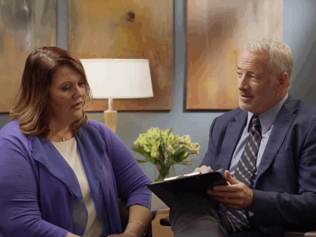 A woman in a purple cardigan sits in consultation with a male doctor holding a clipboard, both engaged in a thoughtful conversation in a warm, professional setting.