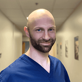 Man with a bald head and beard wearing navy blue medical scrubs, standing in a well-lit hallway with framed artwork.